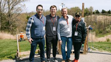 Four Merus employee-owners posing outdoors at an event, with fields and activity stations visible in the background.