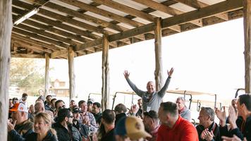 All Merus employee-owners at an annual celebration with one standing up and smiling with hands raised.