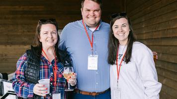 Three people smiling and posing together at an indoor event, wearing name badges and holding drinks.