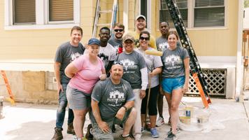 A group of volunteers posing in front of a house they are painting, with ladders and painting supplies in the background.