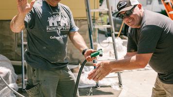 Two volunteers posing by a house they are painting, with ladders and painting supplies in the background