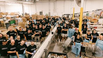 A large group of volunteers wearing matching shirts, working together in a warehouse filled with boxes and food items