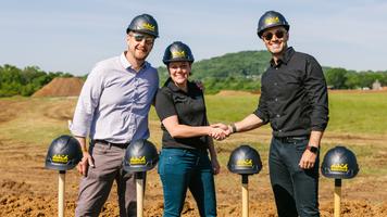 Three people wearing hard hats at a groundbreaking ceremony, shaking hands with shovels in the ground and a scenic landscape in the background.