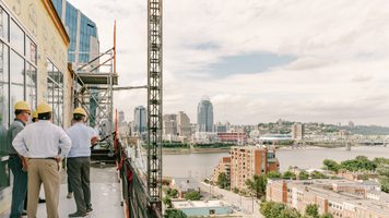 People on a hard hat tour standing on a building under construction, overlooking a cityscape and river,