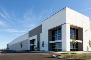Industrial warehouse with white and gray exterior, large windows, and small trees lining the sidewalk