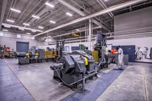 Industrial facility interior with large machinery, high ceilings, and workers in protective gear.
