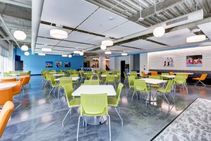 Bright office cafeteria with green and orange chairs, white tables, and colorful wall decor.