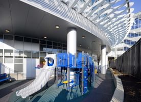 Outdoor childrens play area with blue equipment under a modern curved canopy structure.