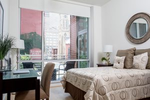 Bedroom with large windows, desk, neutral decor, and city views in the background.