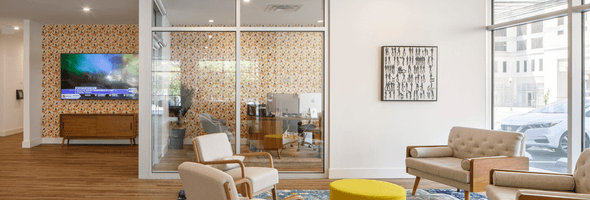 Tenant lounge with patterned wallpaper in background, beige chairs, yellow ottoman, and a glass-enclosed office space.