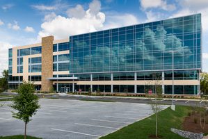 Modern office building with large glass windows, stone accents, and a parking lot under a blue sky.