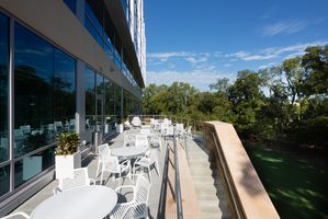 Outdoor tenant lounge beside an office building with white tables and chairs. A staircase and green trees are adjacent.