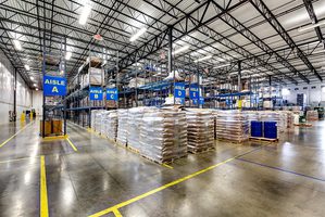 Warehouse interior with stacked pallets, labeled aisles, and high industrial shelving under bright lighting.