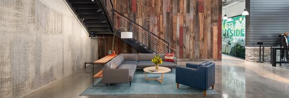 Lobby with a gray sectional, blue chair, wood accents, and rustic wood panel walls under stairs.