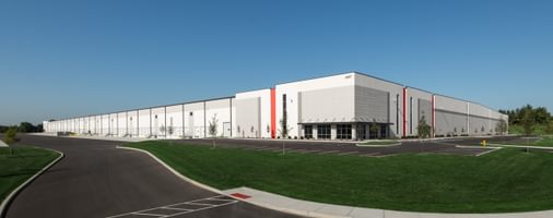 Wide-angle view of a large white warehouse with red accents, lush green lawn, and ample parking space.