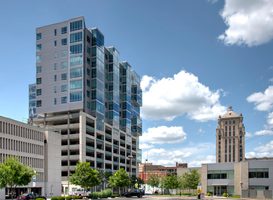 Apartment tower with glass windows and a parking garage below, set against a blue sky and cityscape.