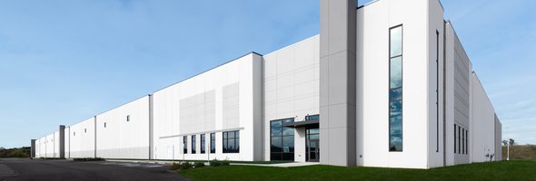 White industrial building with tall glass windows, gray accents, green grass, and a clear blue sky.