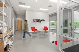 Office lobby with red chairs, Standard Textile logo on wall, and folded textiles on shelves. A person is walking by.