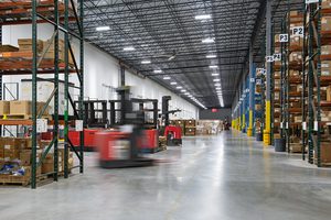 Warehouse interior with tall racks, stacked boxes, forklifts in motion, and bright overhead lighting.